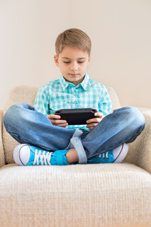 Boy playing hand-held video game on sofa at homeの写真素材