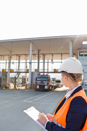 Female worker writing on clipboard while looking at trucks entering in shipping yardの写真素材
