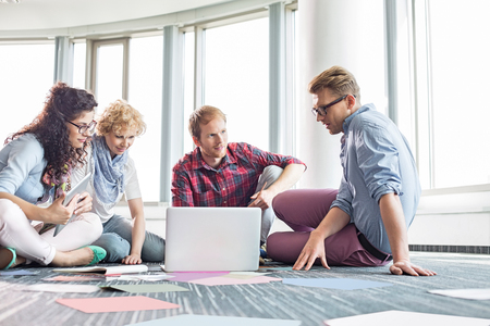 Businesspeople using laptop while sitting on floor at creative officeの写真素材