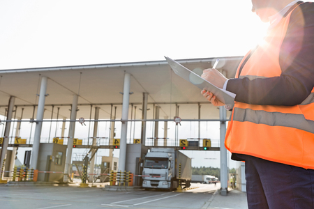 Midsection of female worker writing on clipboard while looking at truck entering in shipping yardの写真素材