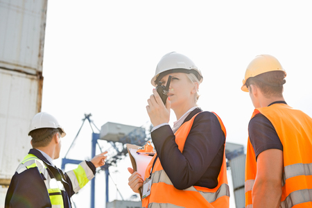 Female supervisor using walkie-talkie while workers discussing in background at shipyardの写真素材