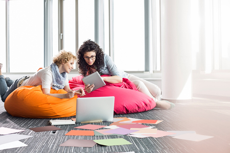 Creative businesswomen using tablet PC while relaxing on beanbag chairs at officeの写真素材