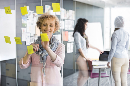 Businesswoman reading sticky notes on glass with colleagues in backgroundの写真素材