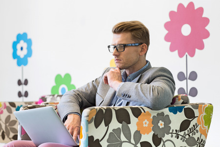 Mid-adult businessman using laptop on armchair in creative officeの写真素材