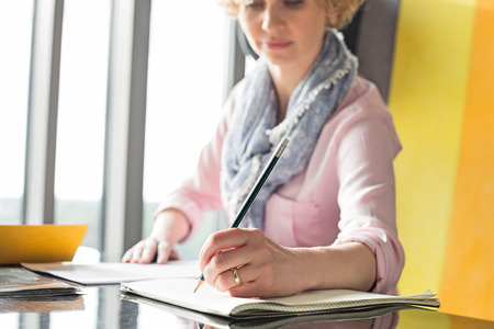 Mid adult businesswoman writing on notepad at desk in officeの写真素材