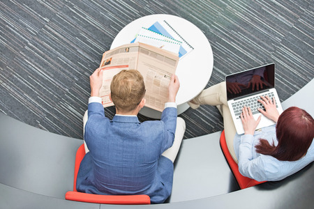 High angle view of businessman reading newspaper while female colleague using laptop in officeの写真素材