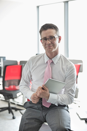Portrait of confident businessman with tablet PC sitting on desk in officeの写真素材