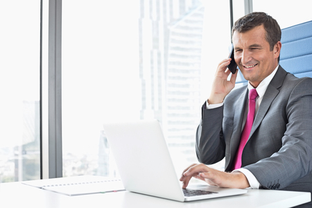Smiling mature businessman talking on cell phone while using laptop in officeの写真素材