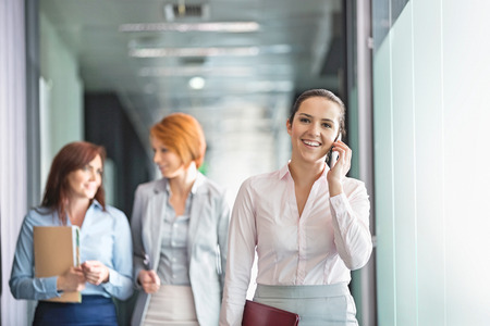 Young businesswoman on call with colleagues in background at office corridorの写真素材