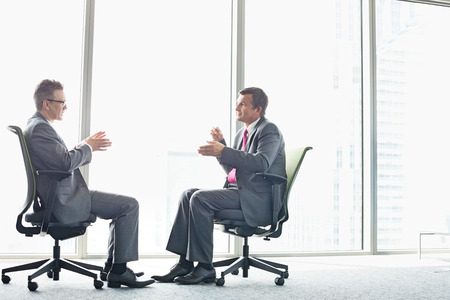 Full-length side view of businessmen discussing while sitting on office chairs by windowの写真素材