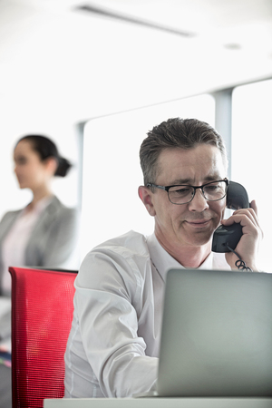 Businessman talking on telephone with colleague in background at officeの写真素材