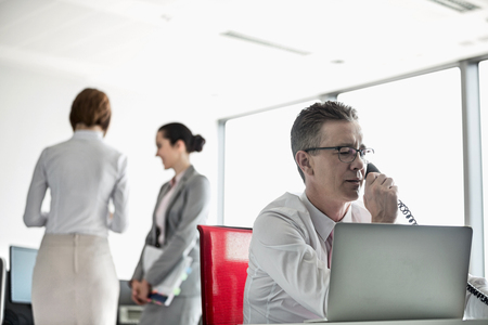Businessman talking on telephone with colleagues in background at officeの写真素材