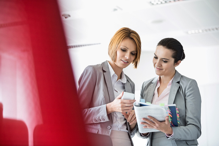 Young businesswomen with book while using cell phone in officeの写真素材