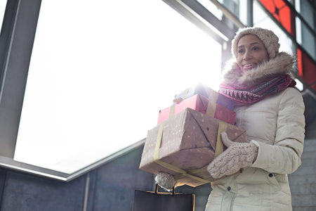 Smiling woman in warm clothing carrying stacked gifts by windowの写真素材