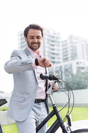 Happy businessman with bicycle checking time outdoorsの写真素材