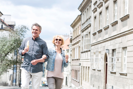 Happy middle-aged couple holding ice cream cones while walking in cityの写真素材