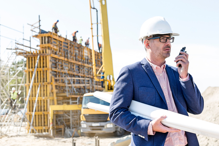 Architect using walkie-talkie while holding blueprints at construction siteの写真素材