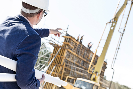 Rear view of architect holding blueprints while pointing at construction siteの写真素材