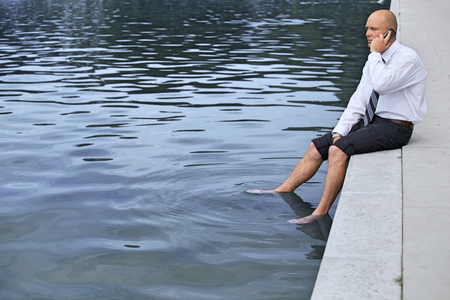 Businessman sitting on wall by lake, using mobile phoneの写真素材