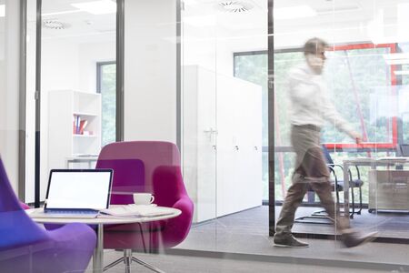 Laptop on table by pink chairs with defocused businessman walking in backgroundの写真素材