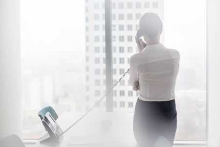 Rear view of businesswoman talking on landline phone while standing at officeの写真素材