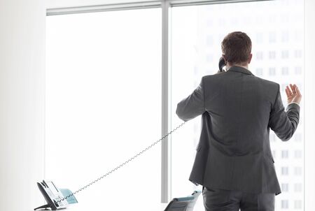Rear view of businessman talking on telephone while looking through window in officeの写真素材