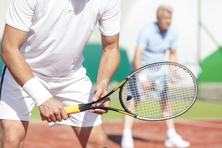 Midsection of man standing with tennis racket against friend playing doubles match on courtの写真素材