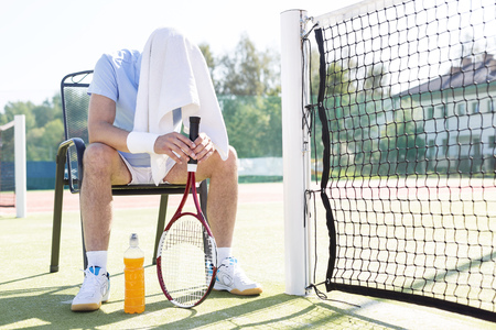 Full length of tired mature man with covered head sitting on chair by net at tennis court on sunny dayの写真素材
