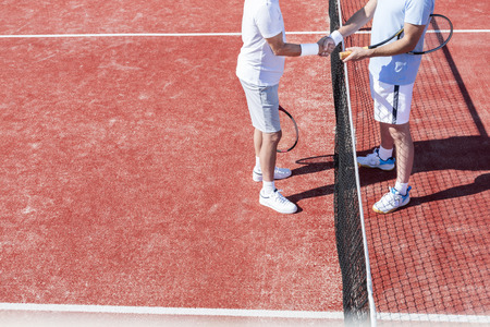 Low section of men shaking hands while standing by tennis net on red court during matchの写真素材