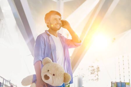 Portrait of man talking on smartphone while holding a teddy bear present for his daughter in airportの写真素材