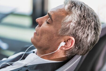Portrait of mature businessman listening to music on his smartphone while sleeping and waiting for boarding in airportの写真素材