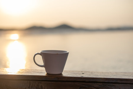 A cup of coffee on the wooden deck, with sunrise and sea view in the backgroundの写真素材