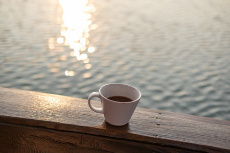 A cup of coffee on the wooden deck, with sunrise and sea view in the backgroundの写真素材