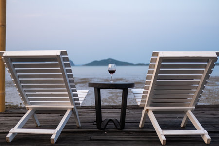 A glass of red wine on the table, with two white benches, beach backgroundの写真素材