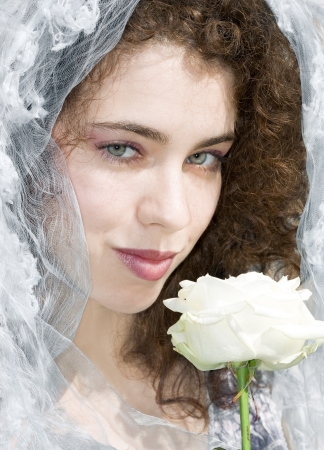 Attractive young woman with a bridal veil is smiling at the camera and smelling a white rose. Wedding and celebration concept.の写真素材
