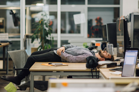 Overworked young man sitting at workplace and sleeping, napping at officeの素材
