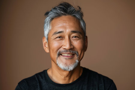 Mature chinese man with gray hair smiling and looking at camera against brown background in studio shot.の素材