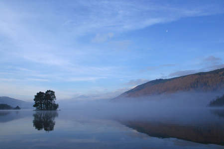 A misty Loch Tay with a small, forested island and the Moon in the skyの写真素材