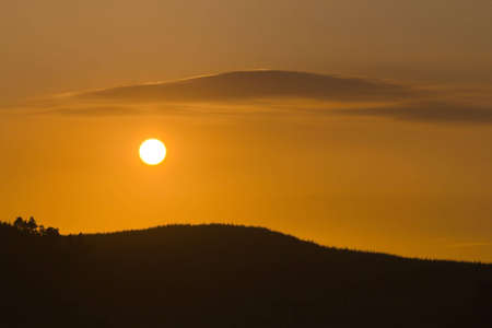 Setting Sun between forested hill and a cloud that mirrors its shape