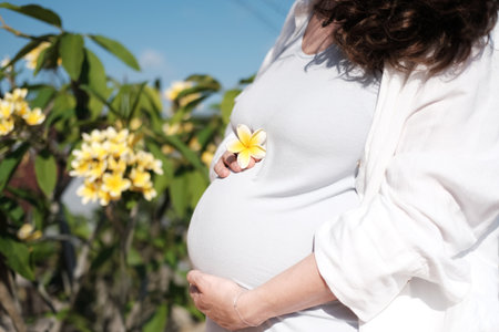 A young pregnant woman stands on a balcony overlooking a garden with a tree with flowers and strokes her belly. Stroking the tummy in which the baby is waiting to be born. Happy motherhood.の写真素材