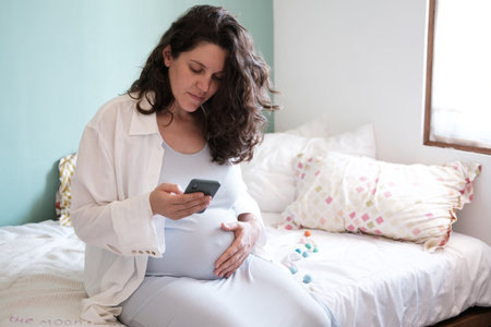 A pregnant woman with a big belly sits on the bed and holds a phone in her hands. Mobile application for pregnant women. A woman looks at how much time is left until the birth in a mobile app.の写真素材