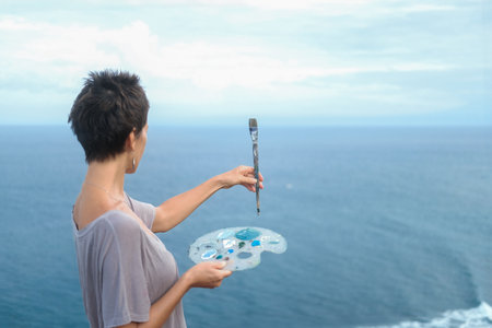 Portrait of an artist against the backdrop of the ocean, who extends her hand with a painting brush in front of her and continues to mix paints. The artist paints a landscape on the ocean from life.の写真素材