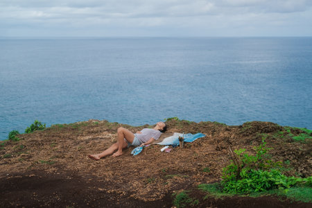 A woman artist lies on the edge of a cliff next to a painting she painted with acrylic paints on canvas. Nature inspiration concept by the ocean. Drawing outdoors.の写真素材