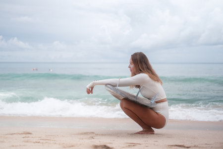 Banner with a young woman in a white surf bikini sitting on the beach with a shortboard and looking forward. Charming sporty female surfer posing on the beach with a surfboard. Water extreme sports.の写真素材