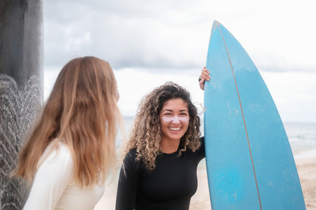 A banner with two young female surfers talking next to their surfboards. Portrait of a Latino woman with curly hair who smiles, her face has colorful zinc and she holds a blue surfboard with her hand.の写真素材