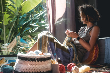 A woman sits on the floor and knits a multicolored blanket from woolen threads in the foreground is a basket with balls of wool in the background is a window behind which there is tropical greeneryの写真素材
