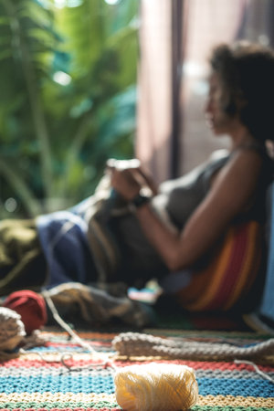 In the foreground is a ball of woolen thread in the background out of focus is a young woman with a short haircut who sits on the floor and knits a blanket with a basket of tools standing next to her.の写真素材