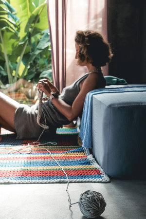 In the foreground is a ball of woolen thread in the background out of focus is a young woman with a short haircut who sits on the floor and knits a blanket with a basket of tools standing next to her.の写真素材