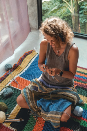 A woman sits on the floor in the bedroom and knits a blanket from woolen threads. The background in the window is tropical greenery. Next to her is a basket with balls of wool and knitting tools.の写真素材