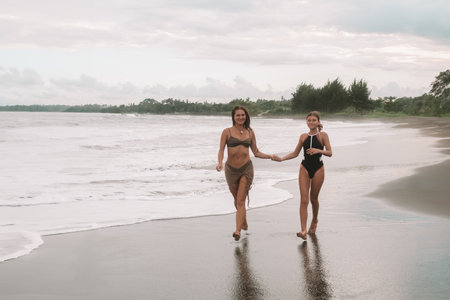 A charming mom and her teenage daughter in swimsuits are running along the black sand beach next to the ocean holding hands and laughing. Vacation with child during the summer holidays. Mothers Day.の写真素材